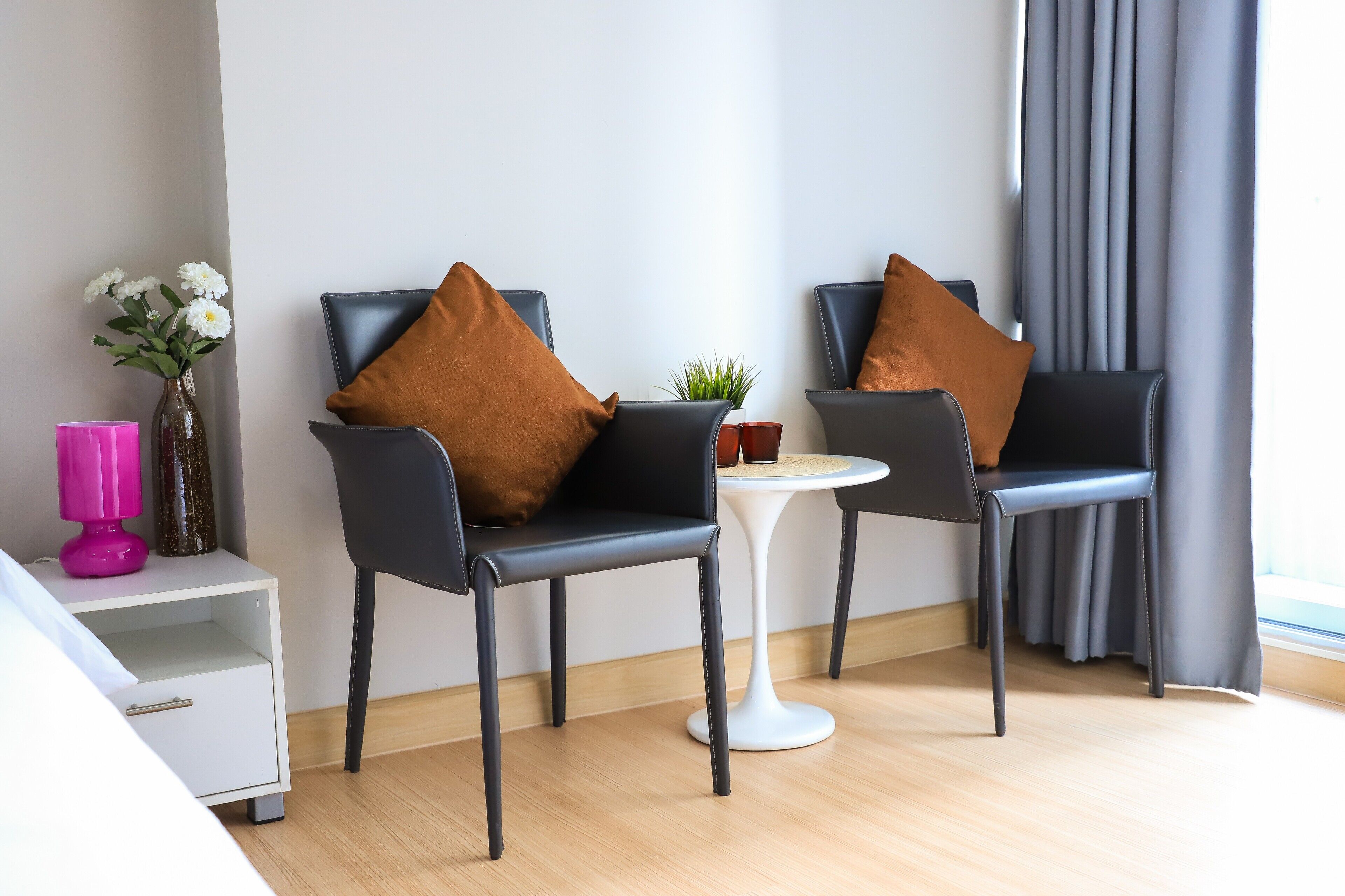 Una habitación de hotel moderna con dos sillones de cuero negro, una mesita blanca, un florero rosa con flores y una mesita de noche blanca.