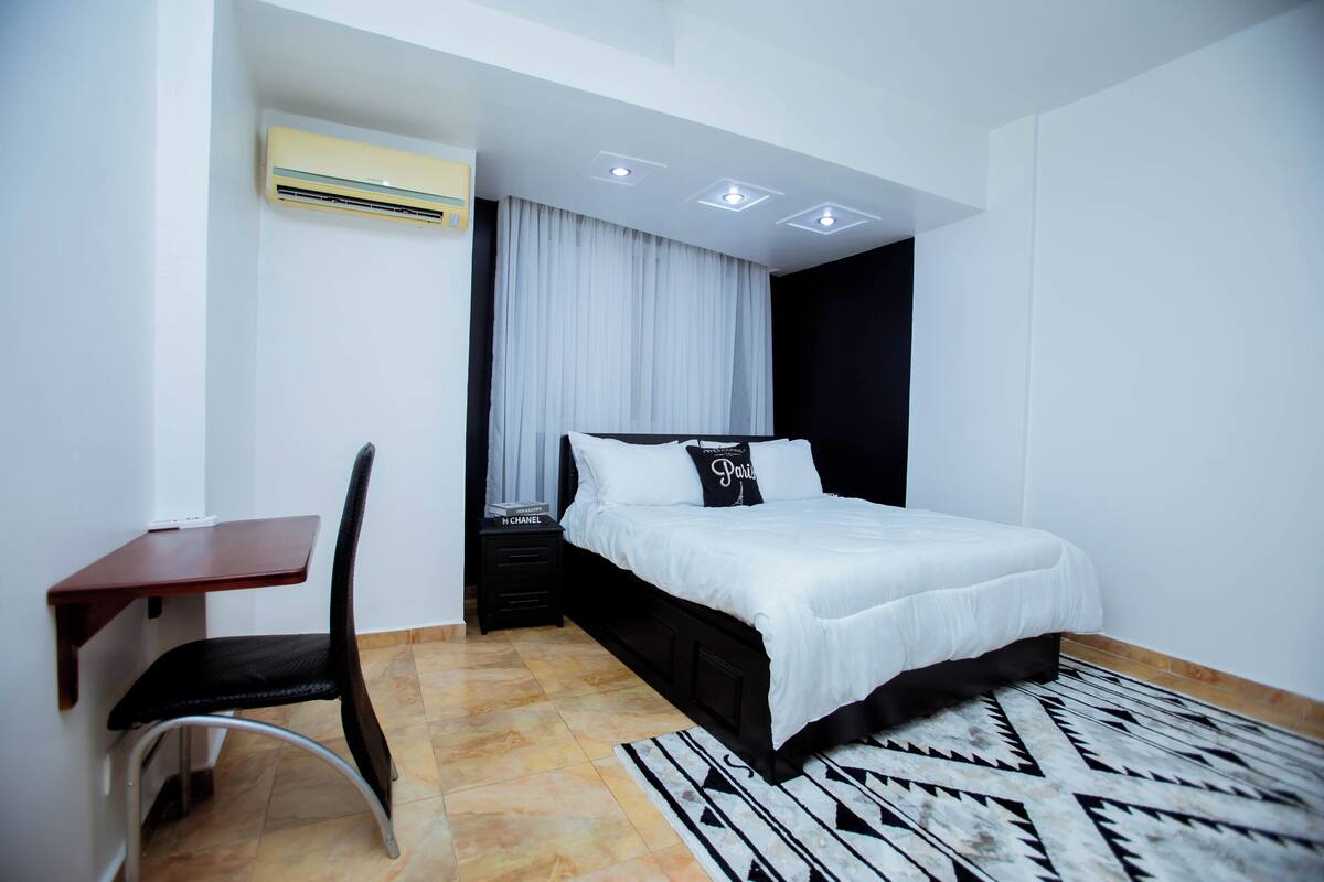 A modern bedroom with a bed, a desk, a chair, and an air conditioning unit.