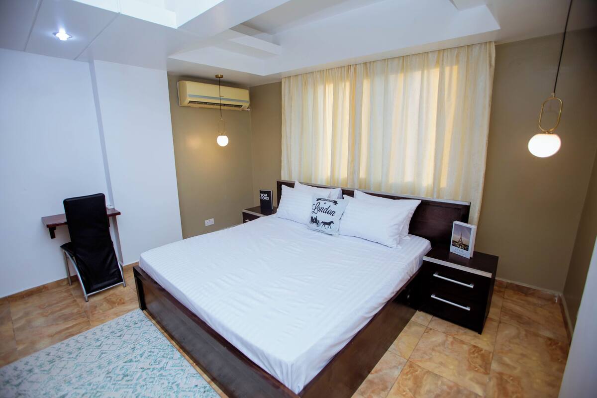 A neatly arranged bedroom with a large bed, a bedside table, a chair, and an air conditioning unit.