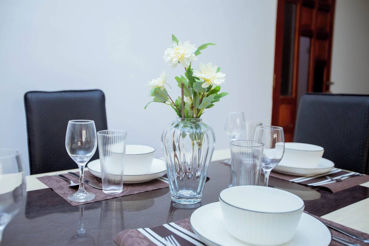 A dining table set with white plates, clear glasses, and a vase with white flowers.