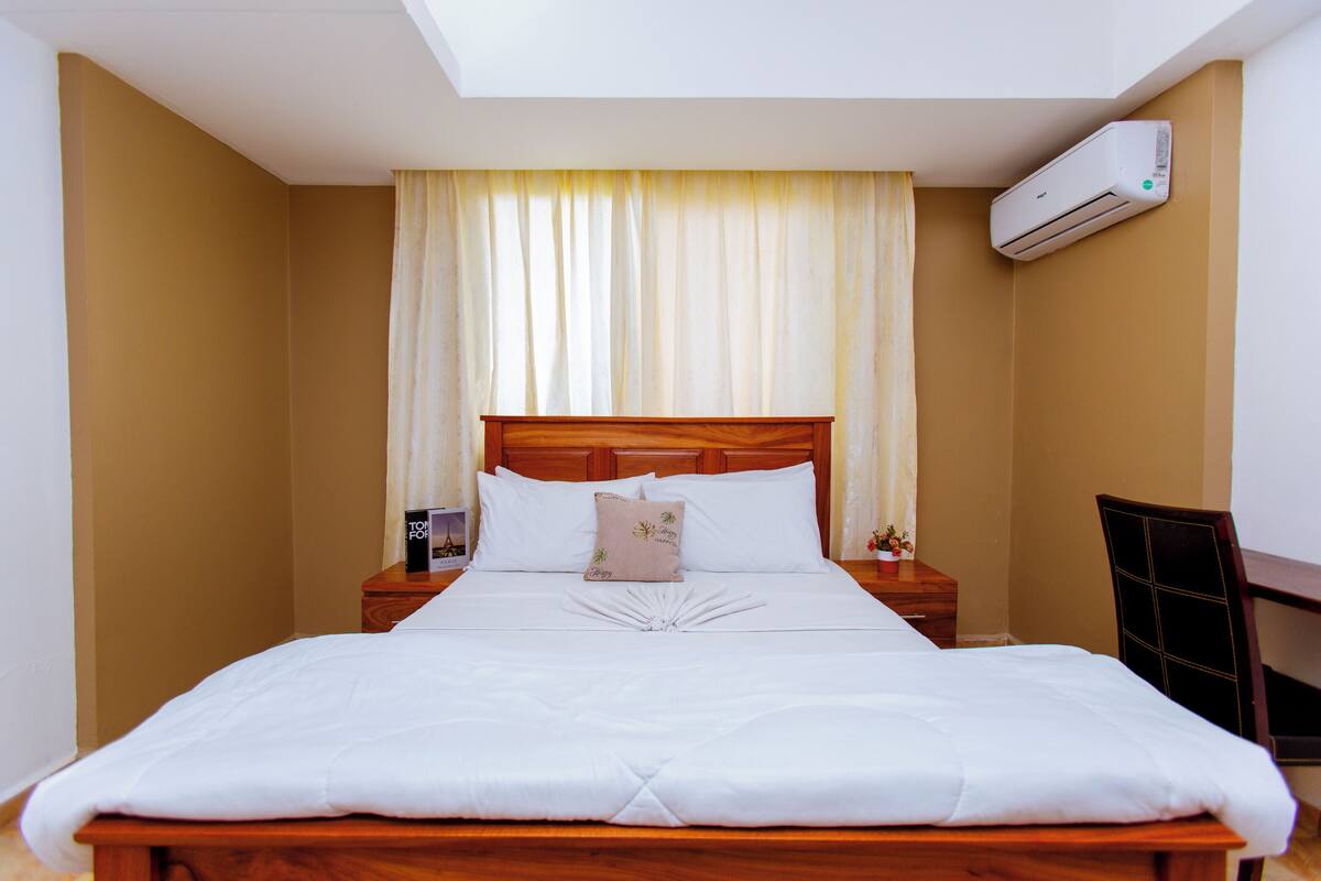A neatly made bed with a wooden headboard, a bedside table with a lamp, and an air conditioning unit on the wall.