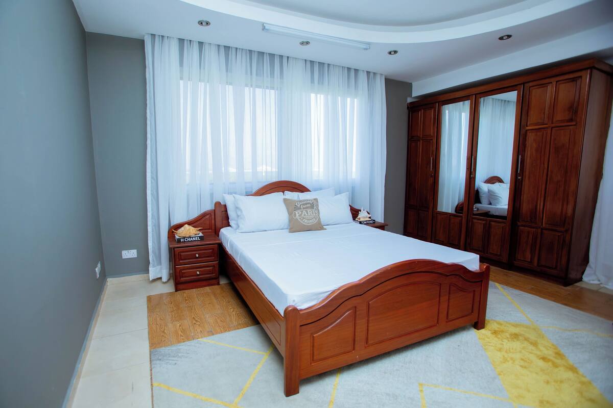 A bedroom with a wooden bed, white bedding, a nightstand, a wardrobe, and a large window with sheer curtains.