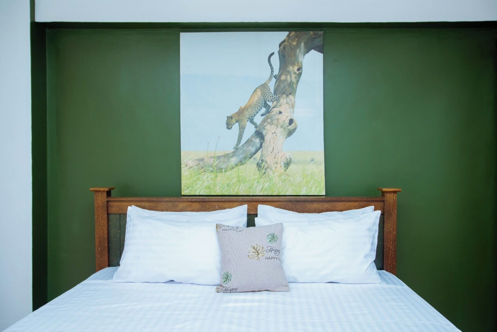 Una cama con sábanas blancas y una sola almohada decorativa, frente a una pared verde con un cuadro de vida silvestre enmarcado encima.