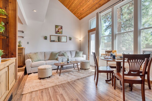 A cozy living room with a wooden ceiling, a white sofa with green cushions, a coffee table, and a dining table with chairs. - 3 Mins to the Lake, Near Tahoe City! Modern Cabin w/ Deck Lounge | Dollar Hill (Tahoe City)