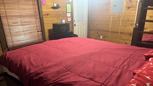 Quarto com piso de madeira, cama com colcha vermelha, pequena mesa preta com micro-ondas e porta com janela.