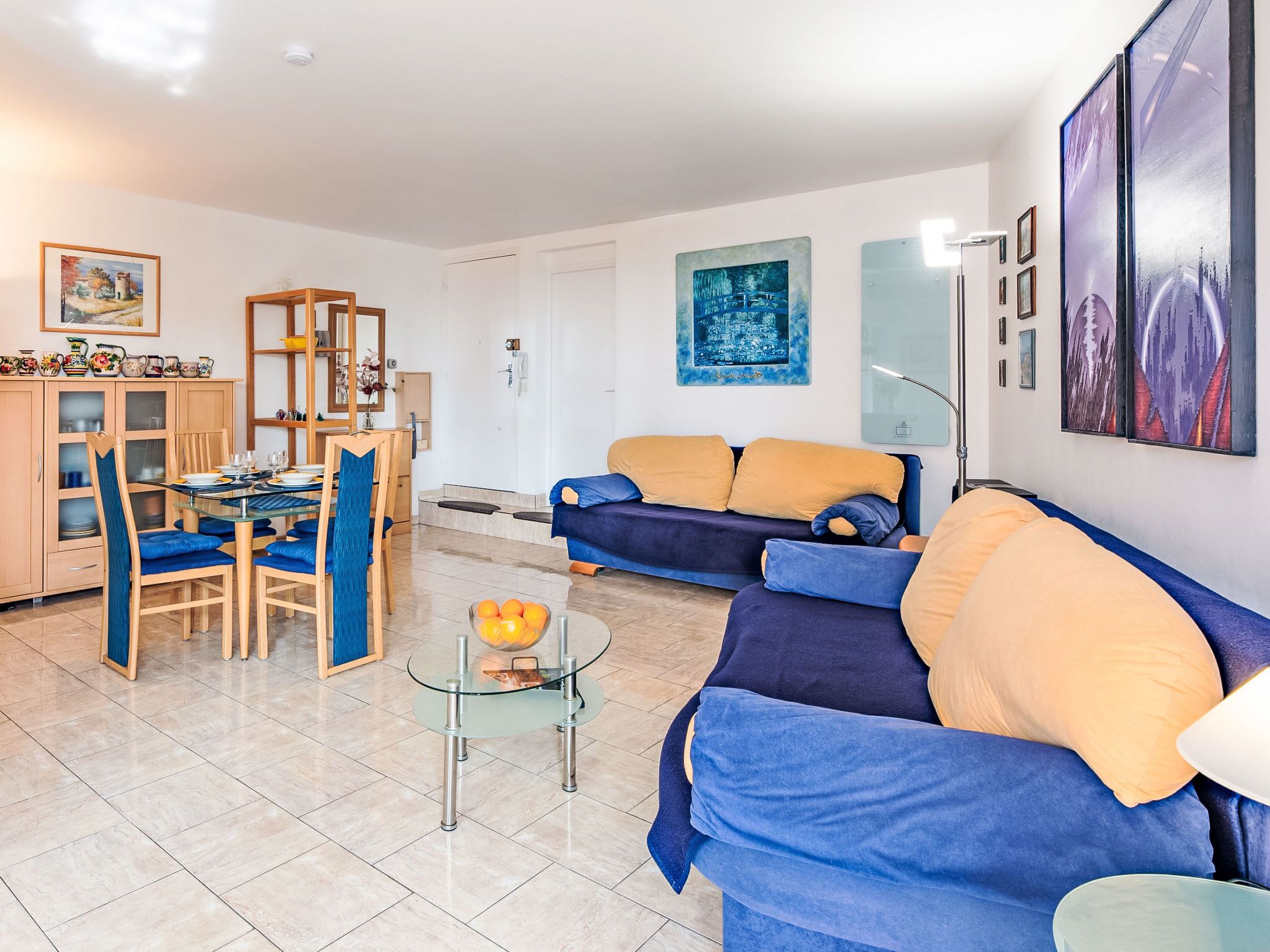 Una sala de estar con un sofá, una mesa de comedor con sillas, una mesa de centro de vidrio y una zona de cocina con armarios y un lavabo.