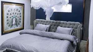 A modern bedroom with a tufted headboard, a large bed with grey bedding, a decorative peacock painting, and a unique chandelier. - Exclusive 4BR/3BA Penthouse Lux Apartment + Rooftop @ BBB Nation Oasis in Kigali (Kigali)