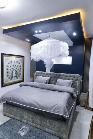 Une chambre à coucher moderne avec une tête de lit capitonnée, un grand lit avec une literie grise, un tableau décoratif représentant un paon et un lustre original.