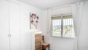 Een kamer met een witte kast, een houten stoel, een schilderij en een raam met uitzicht op gebouwen.