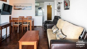 Een woonkamer met een houten tafel, stoelen, een bank en een televisie. Aan de muur hangt een schilderij en er is een keuken in de achtergrond.