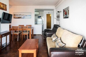 Ein Wohnzimmer mit Holztisch, Stühlen, einer Couch und einem Fernseher. An der Wand hängt ein Bild, und im Hintergrund ist eine Küchenzeile zu sehen.