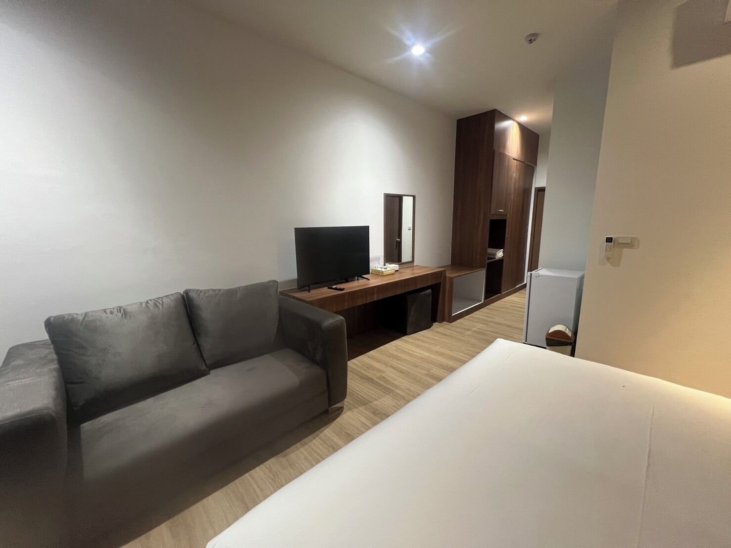 A modern hotel room with a grey sofa, a wooden desk with a TV, and a wardrobe.