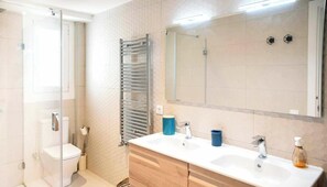 Moderni kylpyhuone, jossa on lasinen suihkuseinä, valkoinen wc-istuin, puinen allaskaappi peilillä sekä suuri peili pesualtaan yläpuolella.