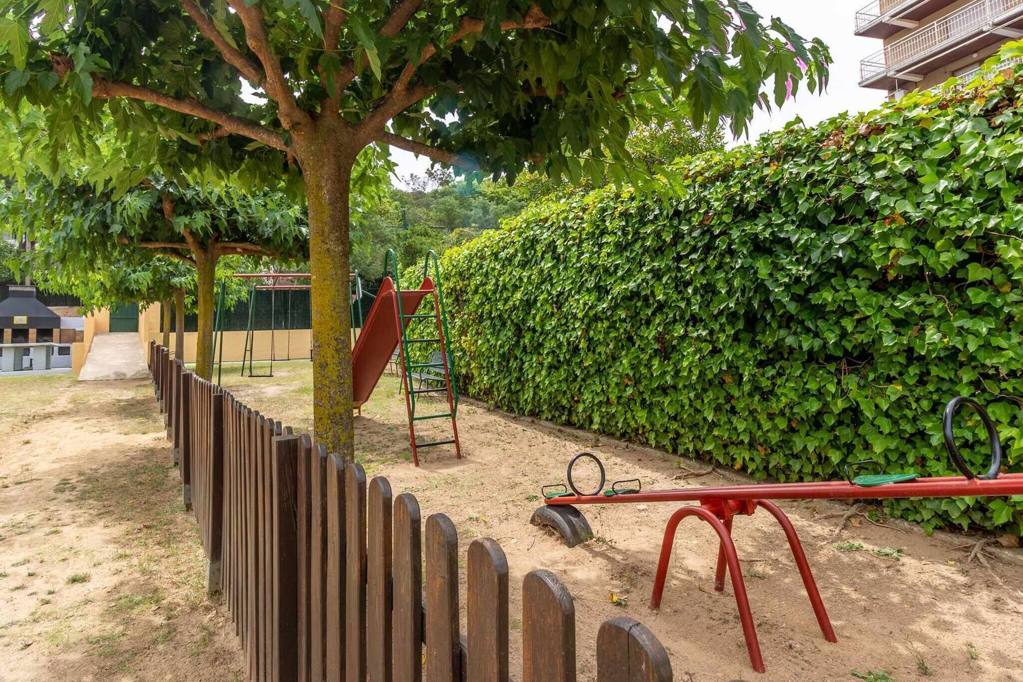 Un parque infantil con un sube y baja, una estructura para escalar y un área cercada con un árbol.