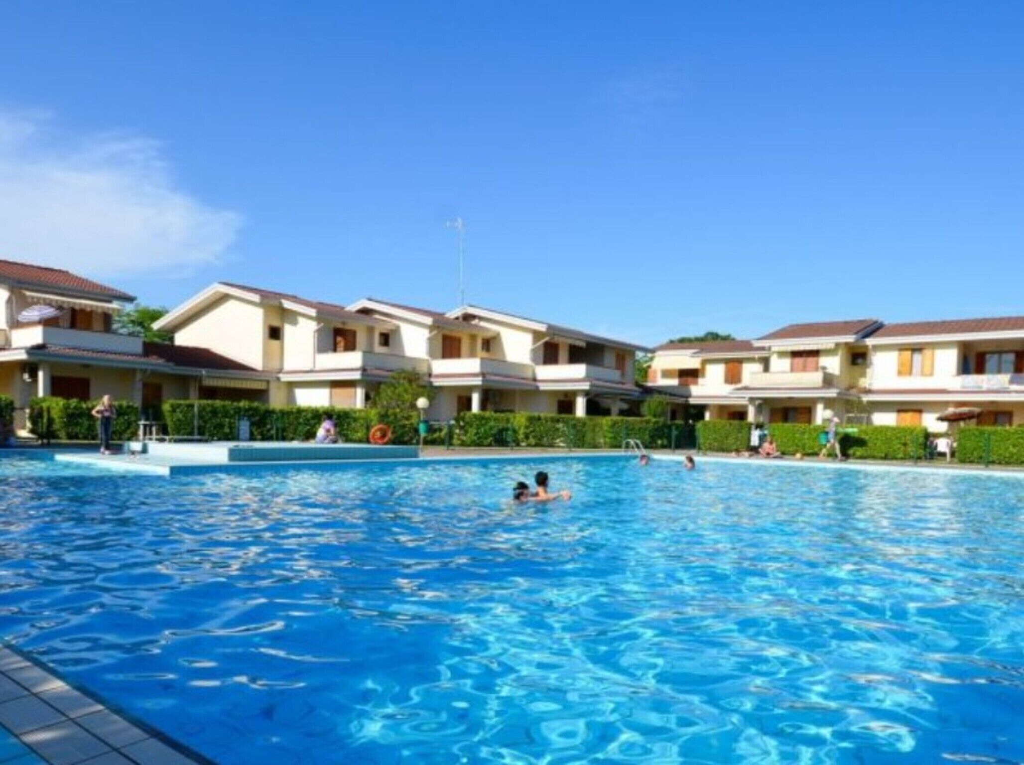 A swimming pool surrounded by multi-story buildings with balconies, clear skies, and a few people swimming.