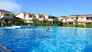 En swimmingpool omgiven av höga byggnader med balkonger, klarblå himmel och några få personer som badar.