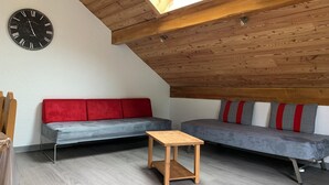 A loft-style room with a sloped ceiling, wooden beams, a skylight, a red and grey sofa, a wooden table, and a wall clock. - 4-person apartment in Pelvoux! 67m2, mountain view Résidence Le Glacier Blanc (Vallouise-Pelvoux)