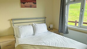 Un lit avec du linge de lit blanc, une table de chevet avec une lampe, une horloge murale et une fenêtre avec des rideaux.