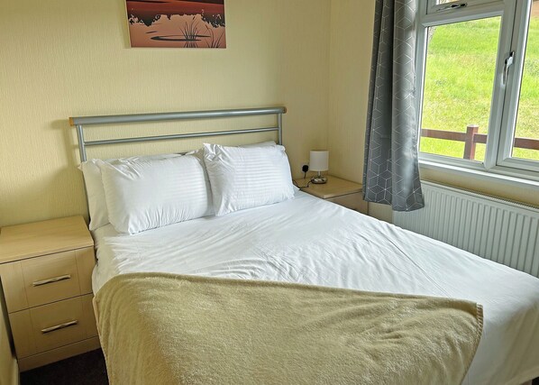 A bed with white linens, a bedside table with a lamp, a wall clock, and a window with curtains.