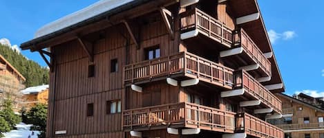 A multi-story wooden building with balconies, a stone base, and a ski slope in the background.