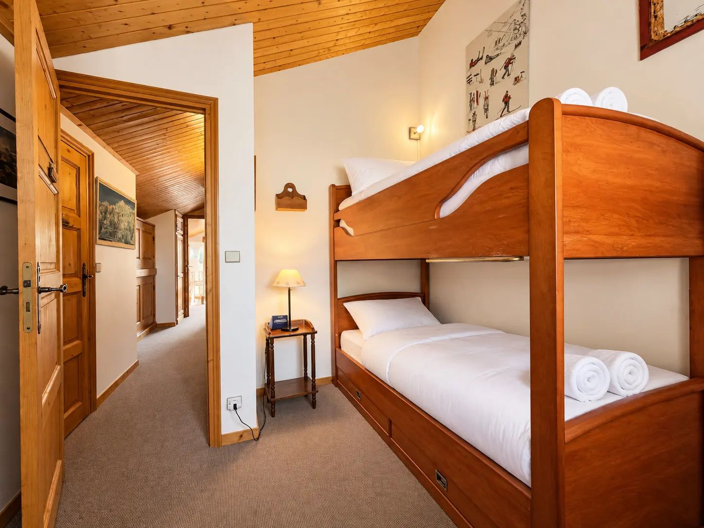 A wooden bunk bed with white bedding and towels, a bedside table with a lamp, and a painting on the wall.