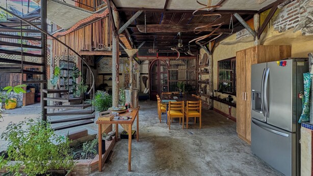 A rustic kitchen with a wooden table, chairs, and a refrigerator. There is a staircase with a rope railing leading to an upper level.