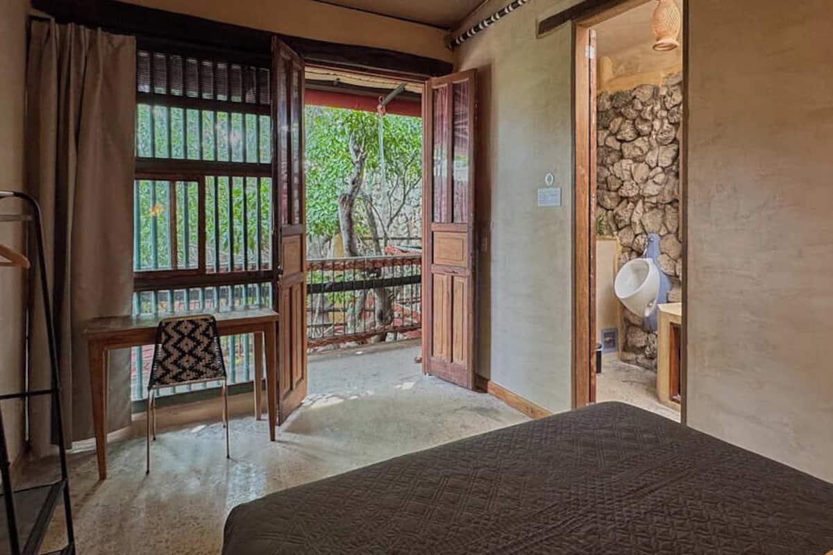 Una habitación con cama, una silla, una mesa, un ventanal con vistas a árboles y una pared de piedra.