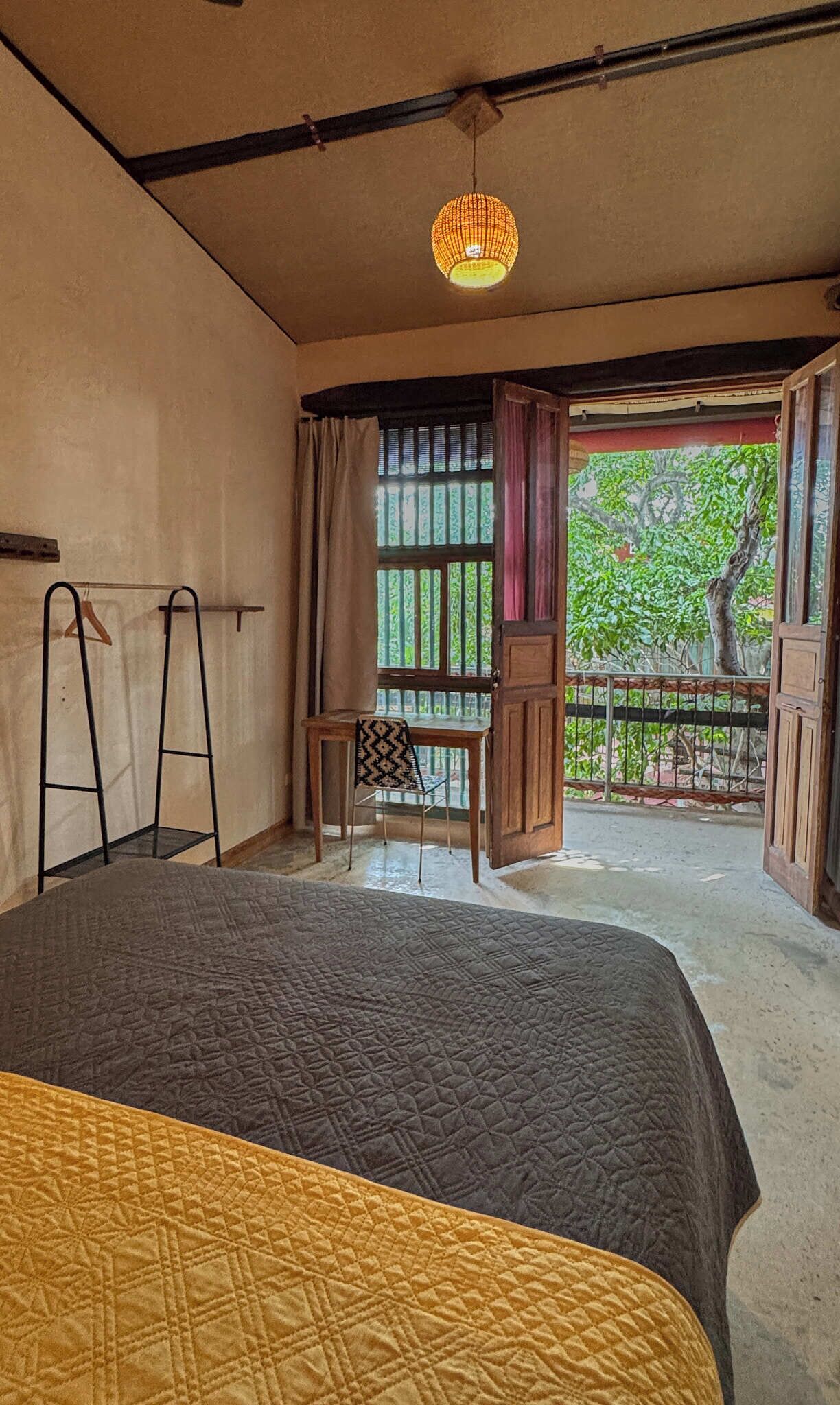 A room with a bed, a desk, a chair, and a view of greenery outside.