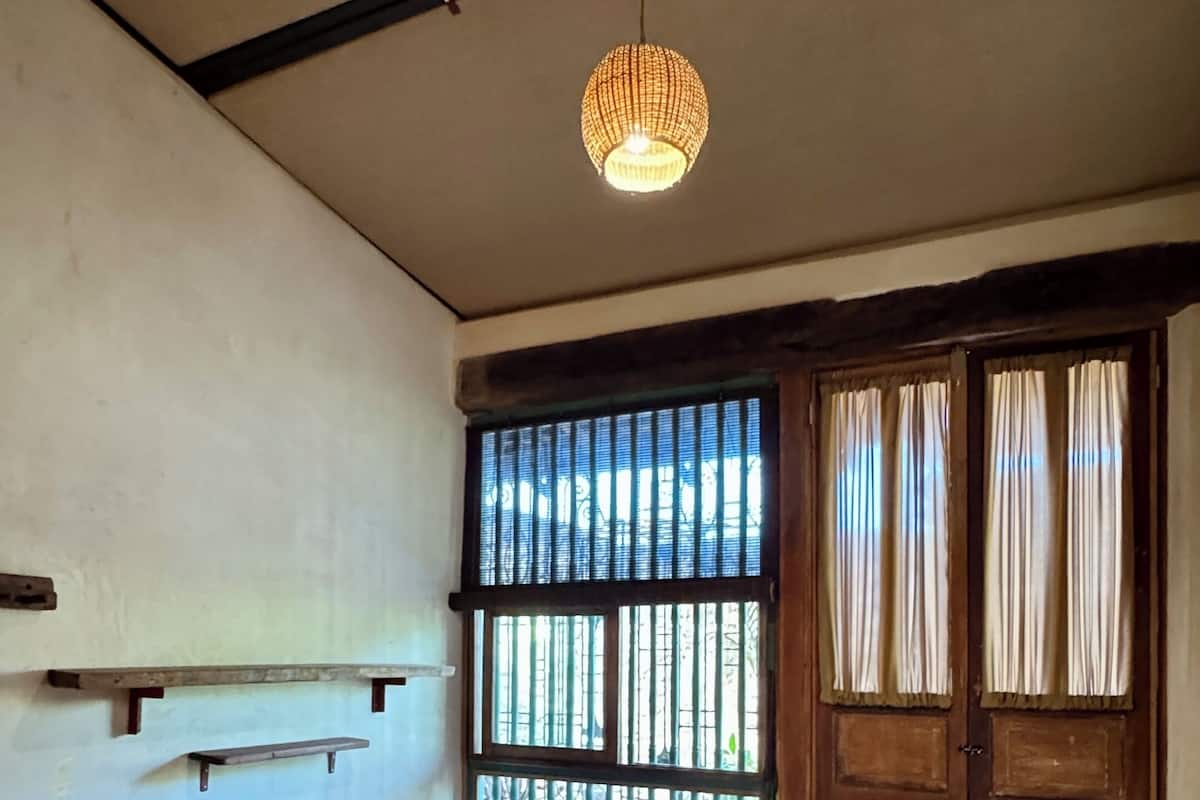Un dormitorio con una cama individual, una lámpara colgante, una ventana con rejas y una puerta de madera.