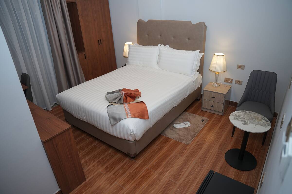 Una cama bien hecha con almohadas y una manta, una mesita de noche con una lámpara, una silla y una pequeña mesa con un control remoto.
