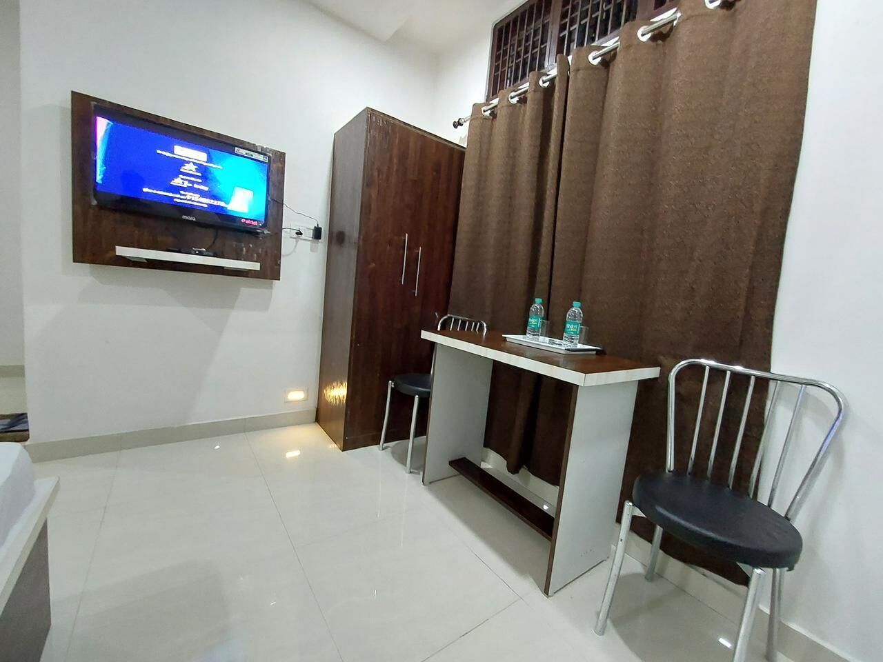 A room with a flat-screen TV mounted on the wall, a wooden wardrobe, a small table with two chairs, and a curtain.