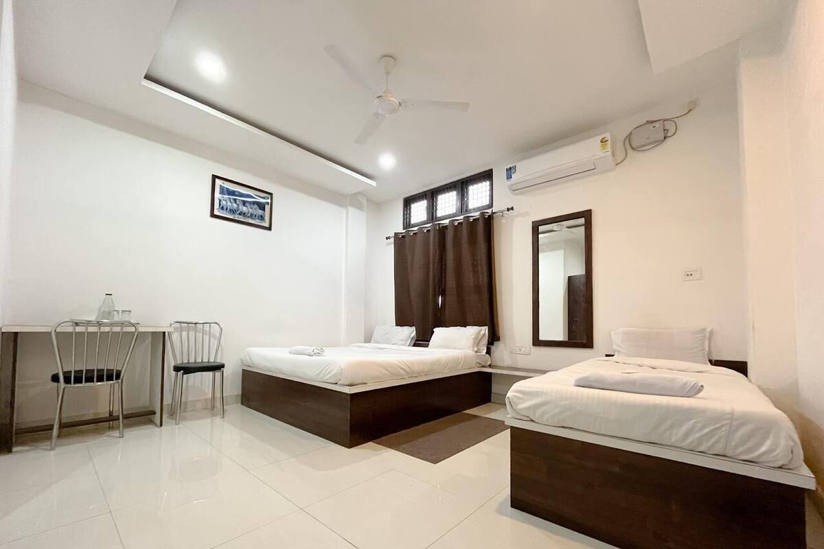 A modern hotel room with two single beds, a desk, chairs, a ceiling fan, and an air conditioning unit.
