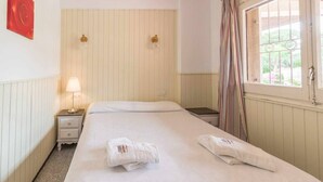 Hotellihuone, jossa on sänky, yöpöytä lampun kanssa, ikkuna verhoilla ja punaisen ruusun kuva seinällä.