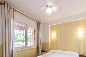 Ett hotellrum med en säng, en takfläkt, ett fönster med gardiner och en väggmonterad lampa.