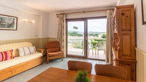 A room with a wooden sofa, a small armchair, a dining table with a potted plant, and a sliding glass door leading to an outdoor balcony with a view of the countryside. - 38.10 S'AgaróCaleta So 206i  Apartment with terrace and views (Sant Feliu de Guixols)