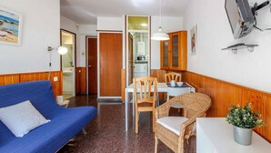 Ein gemütlicher Wohnbereich mit einem blauen Sofa, einem Esstisch mit Stühlen, einer kleinen Küchenzeile und einem an der Wand montierten Fernseher.