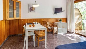 Ein gemütliches Zimmer mit einem Holzkabinett, einem Esstisch mit Stühlen, einer kleinen weißen Ablage, einem Fernseher und einem Fenster mit Vorhängen.