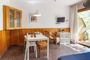 Una habitación acogedora con un armario de madera, una mesa de comedor con sillas, un estante blanco pequeño, un televisor y una ventana con cortinas.