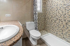 Ein Badezimmer mit einer Granittheke, einem weißen Waschbecken, einer Toilette, einer Dusche mit gemustertem Vorhang und einer gefliesten Wand.