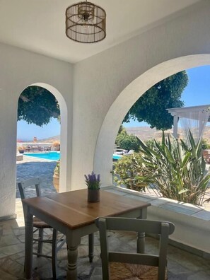 A patio with a wooden table and chairs, a hanging light fixture, and a view of a pool and landscape through arched openings. - Paros Casa Privé (Paros)