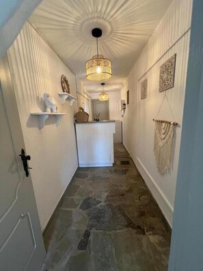 A hallway with a stone floor, white walls, and a central island with a white countertop. There are wall-mounted shelves and a hanging macrame piece. - Paros Casa Privé (Paros)