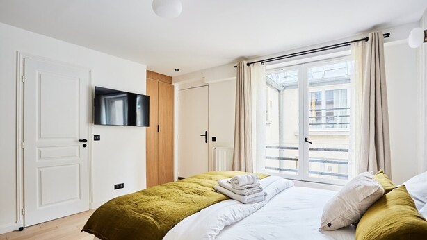 A hotel room with a bed, a television mounted on the wall, a sliding door to a balcony, and a window with curtains.