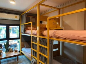 Een slaapzaal met stapelbedden, een klein tafeltje met boeken, een bloempot en een raam met uitzicht.