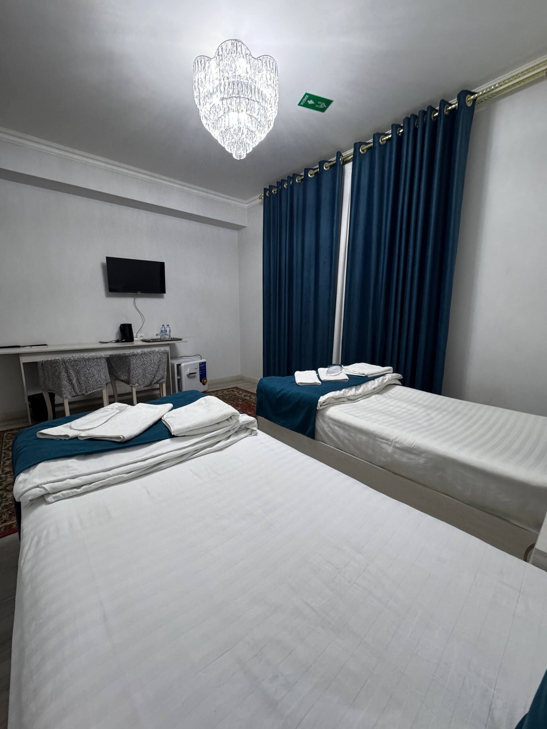 Een hotelkamer met twee eenpersoonsbedden, een bureau met een flatscreen-tv, een kroonluchter en blauwe gordijnen.