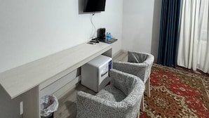 A hotel room with a white desk, a flat-screen TV, two patterned chairs, a small trash bin, and a red patterned carpet. - OKS hotel (Tashkent)