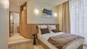 Una habitación de hotel moderna con una cama grande, un panel de madera decorativo, una obra enmarcada sobre la cama y vistas al exterior a través de cortinas translúcidas.
