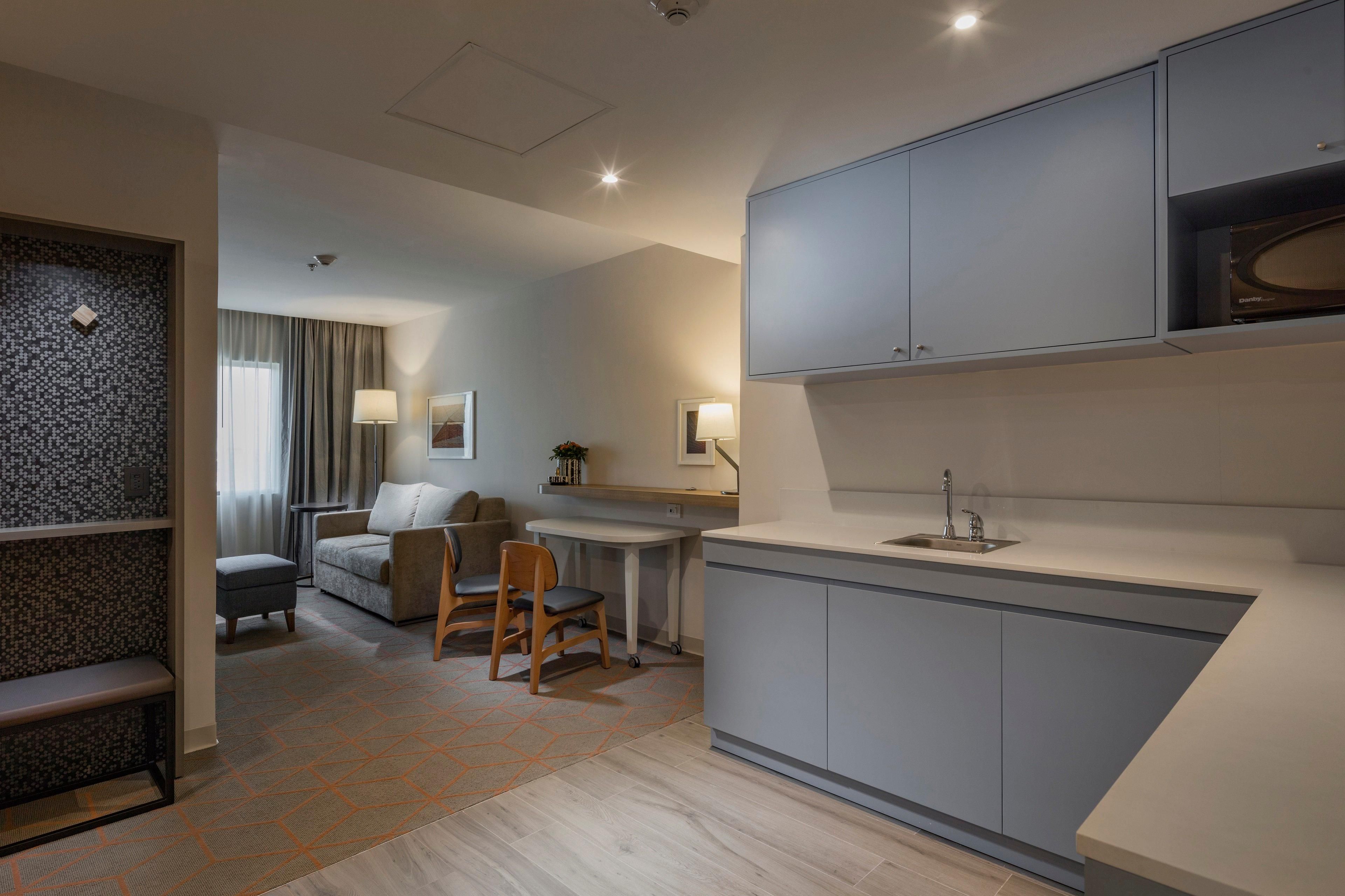 A modern hotel room with a kitchenette, a seating area with a sofa and chairs, and a patterned rug.