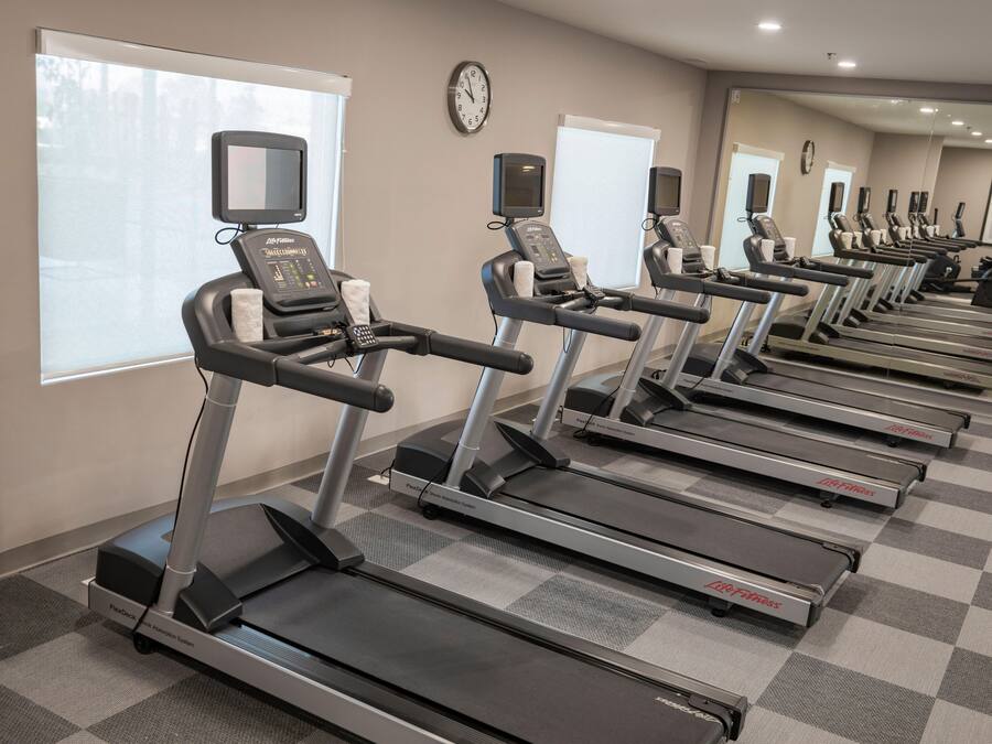 Varias cintas de correr en un gimnasio con suelo de cuadros, un reloj en la pared y ventanas con persianas.