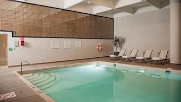 Una piscina interior con suelo de madera, sillones de descanso, un flotador salvavidas y señales de seguridad.