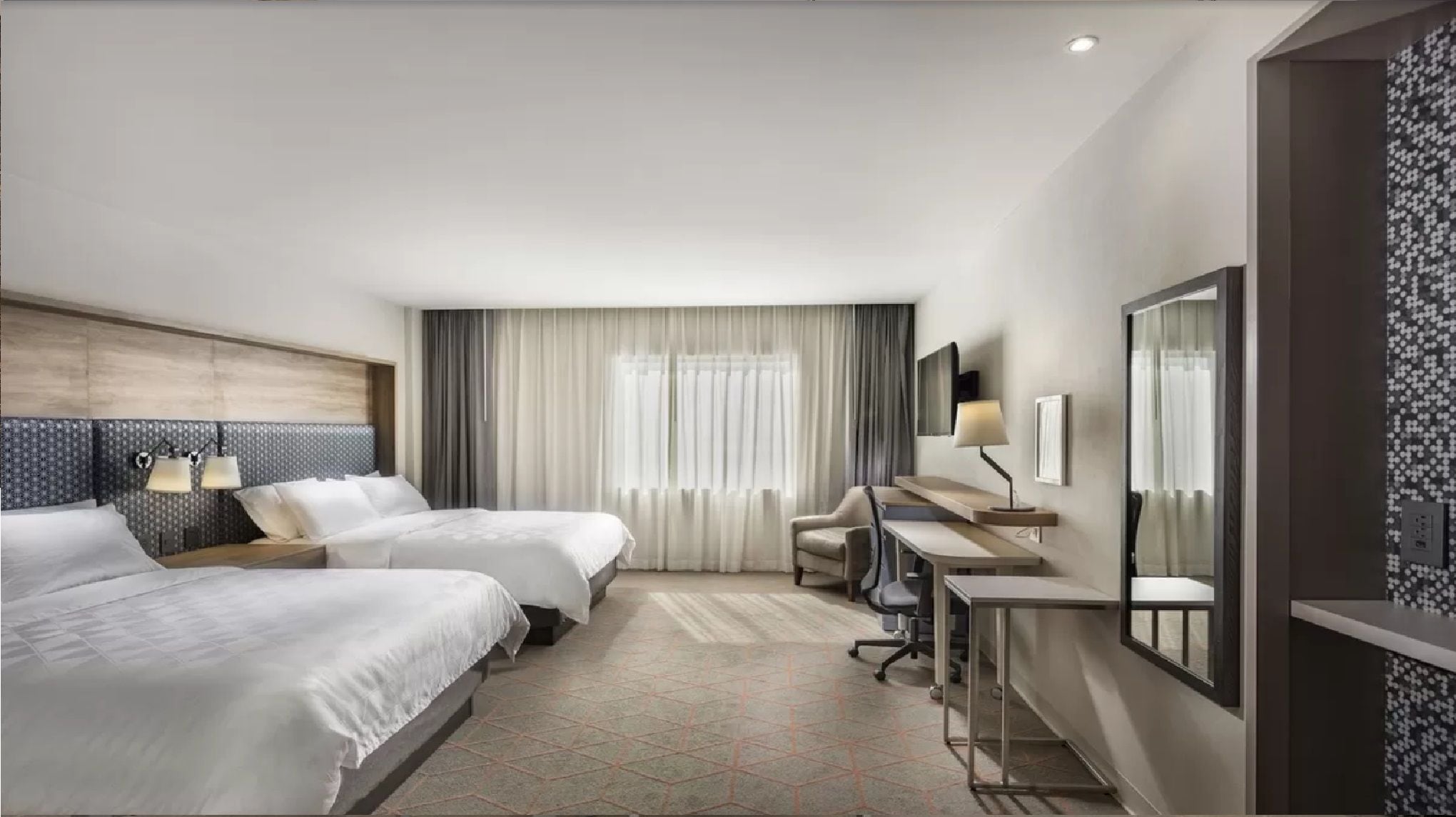 Habitación de hotel con dos camas, un escritorio, una silla, un espejo y una ventana con cortinas.
